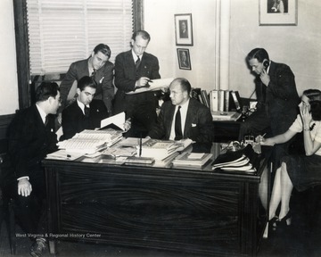Caption on back of photograph reads: "Music and scripts are carefully checked for audience pleasure as J.L. Grimes, general advertising manager for the Wheeling Steel Corporation (in center) and his staff hold a production confab in preparation for the return to the air October 5 of the tuneful "Wheeling Steelmakers" over the NBC-Blue Network." Production staff from left to right: Orchestra leader Tommy Whitley, Maury Longfellow, Ardenne White, Unknown, John Grimes, Unknown, Unknown.