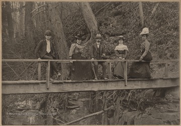 Three unidentified women, accompanied by two unidentified men, are pictured sitting on the railing of the wooden bridge. 