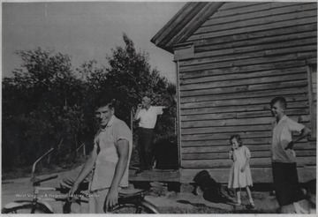 "Buzzy" sits on his bicycle next to two unidentified children who are stranding beside a building. In the background on the porch smoking is Buzzy's father, Ernest Meador. 