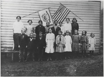 Front row: Foy Meador, Tanner Lilly, Luther Meador, Annie Poff, Eva Hogan Lane Lilly, Minnie Poff, Bertha Hogan Lane, Eva Lilly, Lyda Meadows Neely.Back row: Mary Barker, Sybil Meadows, Mary Lilly Williams, Grover Allen (Betty Allen's brother), Sally Lilly, Eva Meadows, and Arnie Lilly Stoddard.The group poses in front of the West Virginia state flag and the American flag.