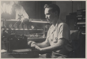 Long pictured typing on a type writer. In the background, an unidentified man is speaking on the telephone.