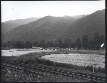 View of the lake, pool, and baseball diamond located beside railroad tracks and a range of mountains. In the background, the building reads, "Bass Lake Park: Free Picnic Tables."