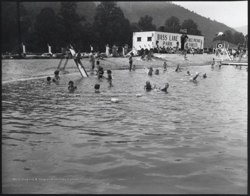 A group of unidentified children swim in the water while parents sun-bathe on the lawn. In the background, a building reads, "Bass Lake Park: Free Picnic Tables".