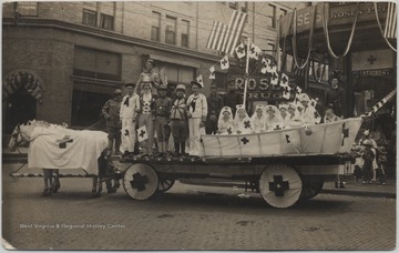 Horses draped in Red Cross flags pull a cart covered in uniformed persons and decorated seats. The drug store is pictured in the background.