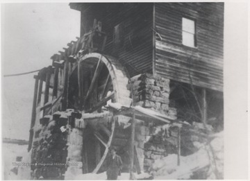 An unidentified man stands in front of the partially frozen water wheel beside the mill building.