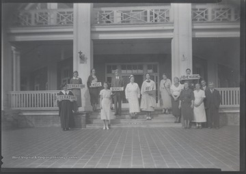 A group of unidentified school teachers and nurse stand in front of the building holding signs that read a variety of school subjects.
