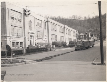 Two school buses sit parked outside of the building. An unidentified student sits on the step railings leading up to the entrance.