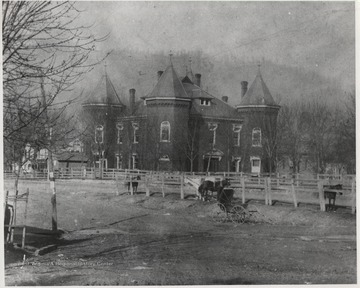 View of the courthouse from across a dirt path. An empty carriage sits beside a small group of horses. 