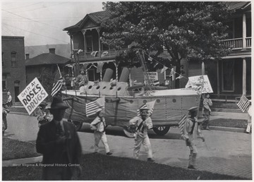 A float shaped as an artillery tank makes its way down Temple Street while young boys dressed as sailors carry American flags and signs for the drugstore. 