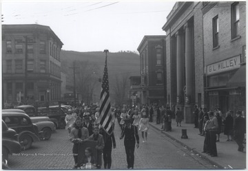 Boy scouts and uniformed military men lead a marching band down the street as citizens watch from the sidewalk.