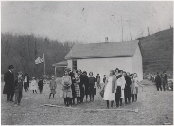 School children gather outside the school building and line up behind a plank of wood. 