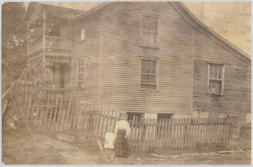 A mother and child stand in front of the fence. A young girl is pictured looking outside the window on the right.
