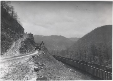 A C. & O. train passes on the right as trucks make travel up the dirt path to aid construction. Workers are pictured in the distance at the top of the dirt ramp. Site located near Sandstone, W. Va.
