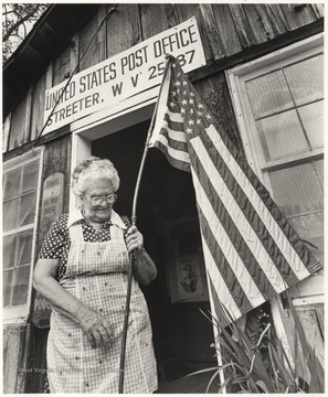 Portrait of the last postmistress at the Streeter Post Office. Pack sets an American flag outside of the building. She served as postmaster since 1949. Her retirement marked the end of the 100-year history of the Streeter post office. 