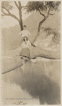 A young woman and man are pictured on top of a large tree branch which hovers over a body of water. Subjects unidentified. 