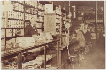 Store clerk L. G. Rhodes pictured behind the counter. Customers in the background are identified as George Collins, Fred Jones (loafer), and Blake Raines (hiding).