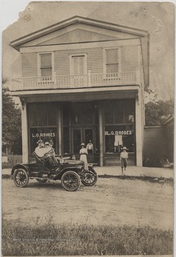 Three men sit outside the store in an old-fashioned automobile while others stand outside the shop. Subjects unidentified. 