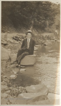An unidentified man sits on a rock beside the water. 