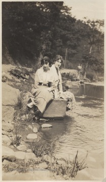 Two unidentified women sit on a rock and dangle their feet above the water. 