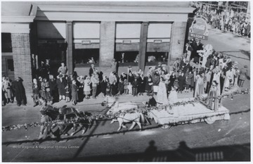 Women dressed in fancy gowns stand on a float as four horses drag it along the corner of Ballengee Street and 2nd Avenue. Spectators line the sidewalks as they observe the spectacle. Subjects unidentified. 