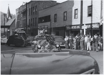 A string of decorated cars make their way through the intersection of Ballengee Street and 2nd Avenue as a crowd watches from the sidelines. 