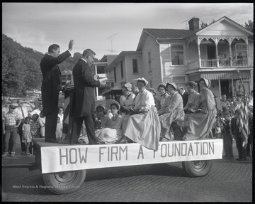 Parade participants dressed in old-fashioned clothing sit on top of a wagon. Attached is a sign that reads, "How Firm a Foundation." Behind the cart, a Boy Scout troop is pictured with an American flag. Subjects unidentified. 