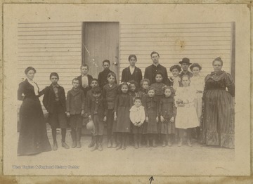Students and faculty pose outside of the schoolhouse for a class photo. The arrow drawn on the card is pointing to Mary Gwinn. Other subjects unidentified. 
