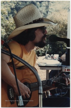 White is pictured playing a guitar while seated behind a microphone. This photograph is from a collection taken at Appalachian Folk Music Festivals, including the Ivydale and John Henry festivals. 