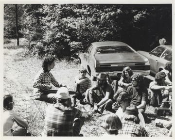 A group sits listening around Edwards, who is playing an instrument. This photograph comes from a series of photos from Appalachian Folk Music Festivals, including the Ivydale and John Henry festivals. 