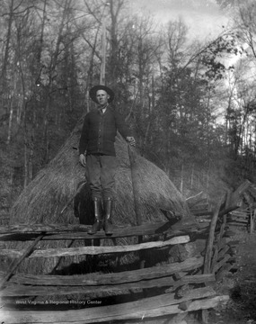 An unidentified man poses in front of a haystack, which is surrounded by a wooden fence. 