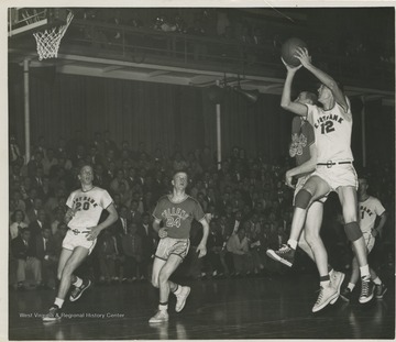 With this shot, West (No. 12) makes the shot that scores him a new state high school record of 860 points in a season with only 6:50 left in the game. Also on the court is Bob Green (No. 20) of East Bank and Bob Short (No. 24) of Mullens.West was East Bank High School's small starting forward. He was named All-State from 1953–56, then All-American in 1956 when he was West Virginia Player of the Year, becoming the state's first high-school player to score more than 900 points in a season.In 1956, West led his team to secure its first ever state championship title. 