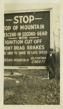 An unidentified woman poses next to a sign for Negro Mountain. Negro Mountain is a part of the Allegheny Mountains that extends from Deep Creak Lake, Maryland to  Pennsylvania. The woman pictured is likely standing next to a sign on Interstate-68 in Maryland.