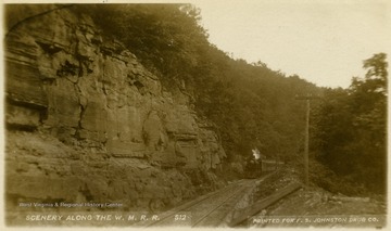 This image is part of the Thompson Family of Canaan Valley Collection. The Thompson family played a large role in the timber industry of Tucker County during the 1800s, and later prospered in the region as farmers, business owners, and prominent members of the Canaan Valley community.A view of the Scenery Along the W.M.R.R. shows a locomotive traveling next to a steep rock face.