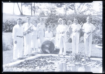 A group of musicians gather around a pond and pose with their instruments. Subjects unidentified. 