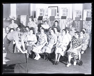 A group of women watch as their instructor demonstrates how to finish furniture. 