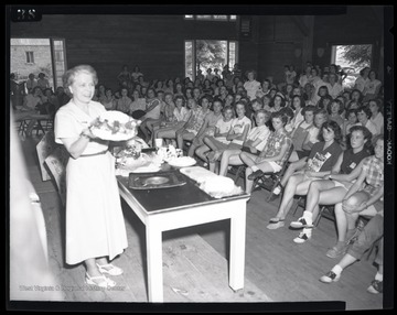 An unidentified woman speaks to a group of young girls as she holds up what appears to be a cake.