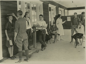 A girl waits as her height is measured on the left, while her companion wait on a scale as her weight is being measured. A nurse in the background points to a series of letters to test eyesight. A young girl sitting on the right waits as her pulse is being taken. Subjects unidentified. 
