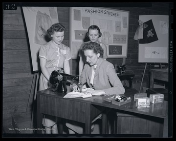 Two girls watch as their instructor demonstrates proper stitching technique. Subjects unidentified. 