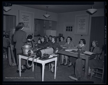 A woman presents vacuum cleaners to a group of women. Subjects unidentified. 