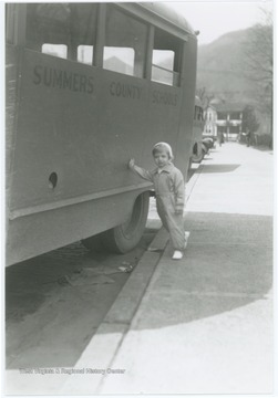 Small boy leans against a school bus owned by Summers County Schools.  A second school bus can be seen in the background.