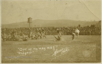 The WVU and W & J football teams play against each other as spectators watch from the sidelines. An inscription on the photo reads, "Out of my way W. & J. -Rodgers", likely referring to the football team captain-elect Errett Rodgers. The Mountaineers won this game 7-0. Subjects unidentified. 