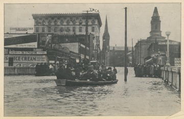 Photo postcard of a flooded street during a 1913 flood in Wheeling, W. Va.  At center, a group of people floats in a boat on the flooded street.  Several signs and buildings are visible in the background, one of which is identified as the Y.M.C.A.  Postcard is part of a souvenir book of 1913 flood images.