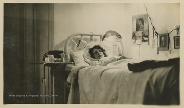 A woman reclines in a bed at Hopemont Sanitarium.  A light fixture is visible, attached to the head of the bed.  Another light fixture is seen plugged into the wall with several photos and items hung on the cord.