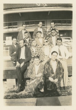 Thirteen men pose in front of a building in various states of dress.  Eleven are wearing bathrobes over lounge clothes, while the other two appear to be fully dressed.  Hospital beds are visible on a porch in the background.