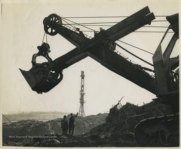 Two coal miners stand beneath the large power shovel machine. Subjects unidentified. 