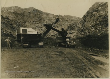 An unidentified coal miner stands beside the power shovel machine as it loads coal into the vehicle. 