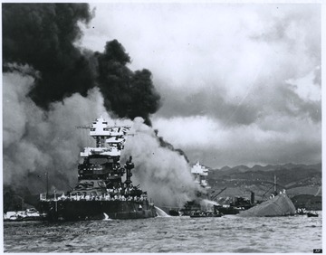 Smoke billows from the U.S.S. West Virginia, which is pictured in the back and center of the photograph. The ship eventually sank.Floating on the left is the U.S.S. Maryland. On the right is a capsized U.S.S. Oklahoma. 