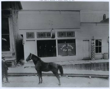 A man leads a horse along the road. Four men stand at the saloon window. 