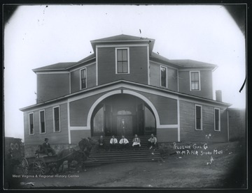 Store manager W. M. Pifer stands in front of the store entrance while his five employees sit on the stairs. An African-American man is pictured in a horse-drawn carriage to the left. 