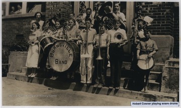 Hobert Cosner is pictured playing the snare drums. A group of students and instructors are holding instruments as they stand on the steps in front of the school building. 
